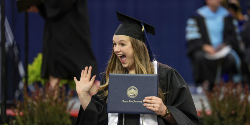 Girl graduate waving and smiling holding diploma.