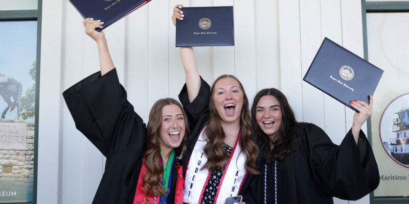 Three girls wearing graduation regalia holding diplomas high overhead and big smiles.