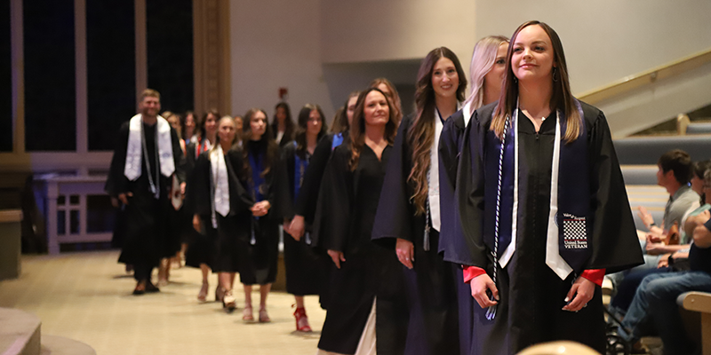 Students walking into auditorium wearing graduation regalia.