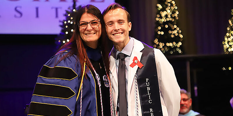 Instructor wearing graduation regalia with student nurse wearing white coat.