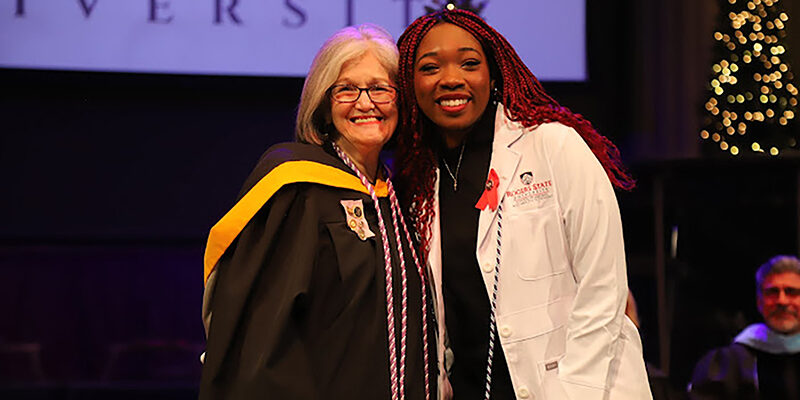 Instructor wearing graduation regalia with student nurse wearing white coat.