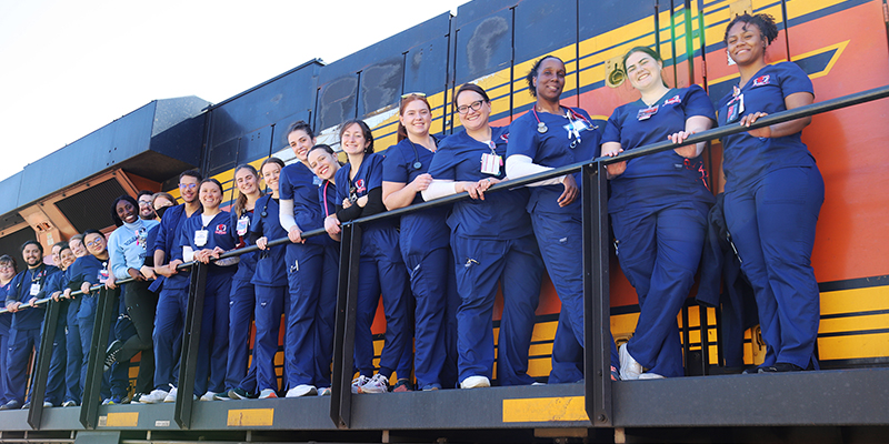 A long row of RSU nursing students, wearing navy blue scrubs, stand long the walkway on a BNSF train engine.